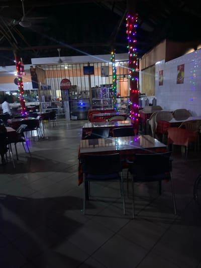 The image captures an outdoor or semi-outdoor dining and gathering area at night, illuminated by numerous colorful string lights. The scene is relatively dim, with artificial lights providing the main illumination.
In the foreground and mid-ground, multiple strands of multi-colored fairy lights are strung horizontally, creating a festive and inviting atmosphere. Below these lights, several tables and chairs are visible on a tiled floor with a diamond pattern, some appearing to be set up for use, others potentially stacked or stored.
In the background, a building or structure with light-colored walls houses what appears to be a commercial establishment, likely a restaurant or cafe. A prominent red circular sign bearing the "Coca-Cola" logo is affixed to one of the walls. Below it, a large glass-fronted refrigerator or display case can be seen, suggesting a beverage or food service area. Other faint posters or decorations are on the walls, but their details and text are not discernible.
A few blurry figures are scattered throughout the space, particularly in the lower left portion of the image, indicating the presence of people. They appear to be engaged in an activity, likely dining or socializing. The time of day is clearly night, evidenced by the darkness and the reliance on artificial lighting for visibility. The overall ambiance is casual and relaxed, fitting for a local eatery or communal space in a location like Bagamoyo, Tanzania.