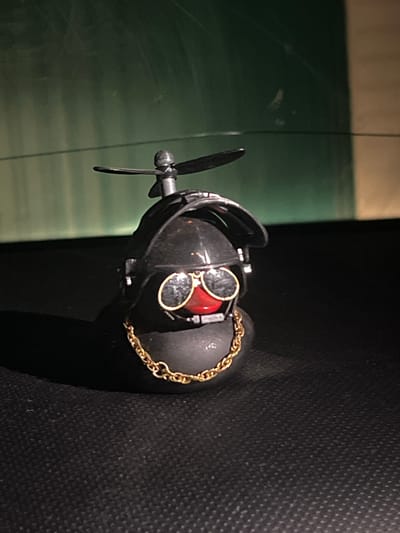 A close-up shows a dark-colored, possibly steampunk-style object featuring a propeller, goggles with red lenses, and a gold chain, resting on what appears to be a dark, textured surface. The object has metallic and black components. A green surface is partially visible in the background. The image doesn't provide information about a location or city.