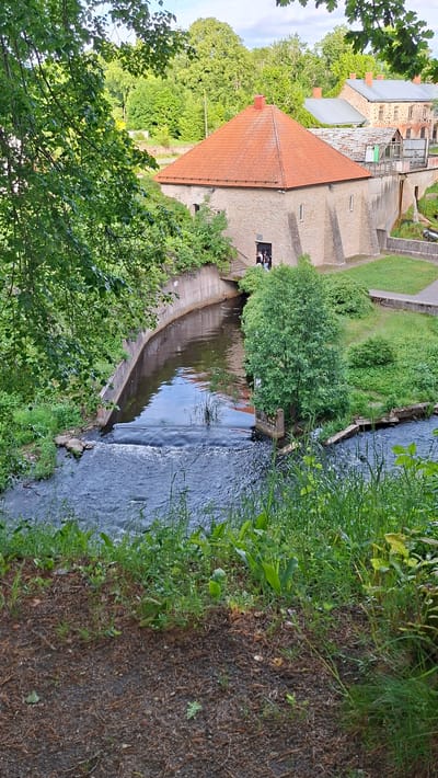 This is a full, eye-level, vertical shot of a canal in Keila-Joa alevik, Estonia, with a building and surrounding greenery. In the foreground, the ground is covered with dirt, pine needles, and sparse grass. A narrow, dark blue canal curves from the upper left to the lower right, bordered by concrete walls on the left and right. A small, shallow waterfall or dam is visible in the canal, creating ripples. Lush green grass and plants grow along the canal banks and the foreground.

The main subject of the midground is a light-colored stone building with a red, pyramidal roof. The building has a prominent chimney and several small, rectangular windows. Two people, one in a light-colored top and dark bottoms, and another in a dark top and light bottoms, are standing on a walkway leading to an entrance on the left side of the building. To the right of the building, there is a grass-covered slope and more vegetation, with a hint of other buildings in the background.

The background is dominated by dense green trees and foliage, suggesting a wooded area surrounding the location. The sky is partly cloudy. The overall impression is a peaceful, natural setting with a historical building.
