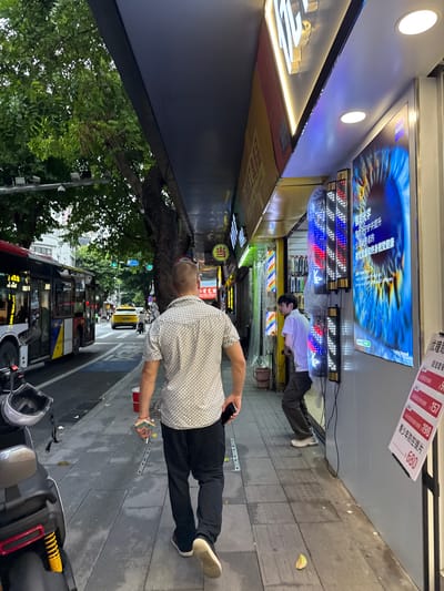 A man walks away from the camera on a sidewalk in Liwan District, China. He is wearing a white patterned short-sleeved shirt and black pants. To his left, a red and white bus drives down the street. Across the street, a yellow taxi is visible, along with motorcycles and other vehicles. The sidewalk is lined with shops displaying illuminated signage and advertisements. One prominent advertisement features a close-up of a human eye. The ground is paved with concrete tiles, and there are trees and streetlights present.