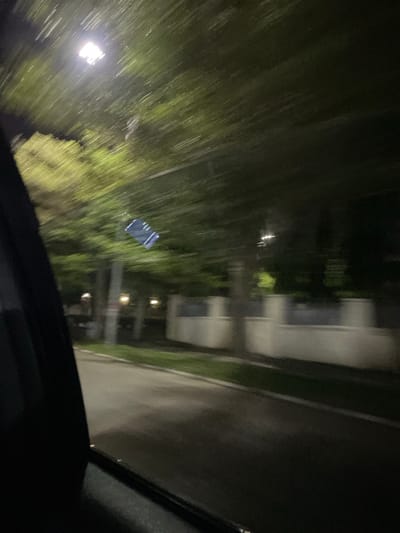 A nighttime, blurred image from inside a moving vehicle. Through the windshield, trees line the road to the left with a streetlight illuminating them. Further along the road is a multi-story building. The windshield wipers are visible on the right side of the frame. It is not possible to discern the exact location in Guzape, Nigeria, or any additional specifics about the environment or any persons present.
