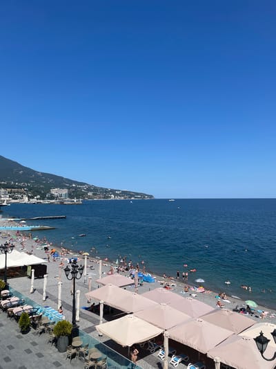 The image depicts a sunny day at a pebble beach resort in Yalta, Ukraine. The foreground features a paved area with outdoor seating, including tables and chairs under tan umbrellas. Beyond this area is a pebble beach populated with numerous sunbathers, many of whom are lying on towels or under umbrellas. People are also seen in the clear blue water, some swimming and others floating on inflatable devices. A pier extends into the sea, and further in the distance, a mountainous coastline with buildings and a lighthouse is visible. The sky is clear and blue.