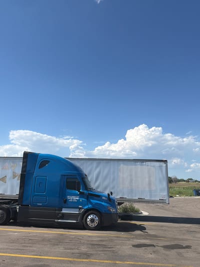 This image depicts a blue semi-truck parked on an asphalt lot in North Platte, United States of America. The truck has "CRETE CARRIER CORP SHAFFER TRUCKING" printed on its side, along with a USDOT number "080071 073705" and "88605" on the front. The truck is positioned in front of a large white trailer, which appears to be connected to another truck partially visible to the left. Yellow lines mark parking spaces on the asphalt. The background features a vast blue sky with scattered white clouds and a distant landscape of dry grass and trees. A second semi-truck, also white, is visible in the far distance to the right. The scene appears to be a rest stop or truck parking area, under a clear, bright sky.
