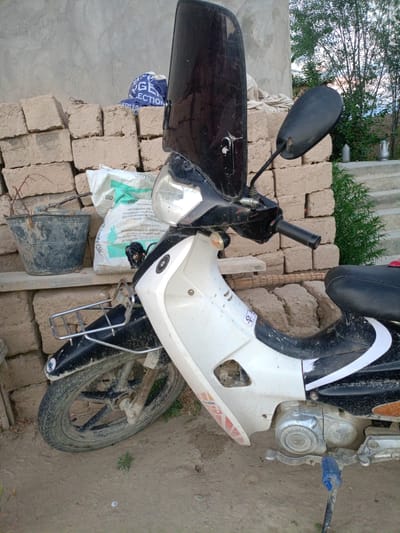 A white and black motorcycle is parked outdoors, leaning against a wall constructed of light-colored bricks. The motorcycle features a large, dark-tinted windscreen. A black bucket containing dried twigs sits to the left of the motorcycle, with a white bag of what appears to be cement or fertilizer behind it. The ground is dirt and appears dry. In the background, there are trees and a concrete structure with stairs. The scene is illuminated by natural light, suggesting it is daytime. The motorcycle is dirty, with mud splattered on its body and wheels.