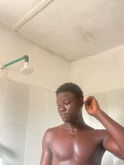 The scene depicts a young adult male in what appears to be a bathroom or shower area in Mosafejaw, Nigeria. The male, with dark skin and short, curly dark hair, is shirtless, revealing his bare chest and shoulders which appear wet or glistening. He is looking downwards and to his right, with his right hand raised near his right ear as if scratching or adjusting something. In the upper left background, a white circular shower head is visible, connected to a green PVC pipe that extends horizontally from a light-colored wall. The wall below the pipe features white tiles. The ceiling above is light-colored and exhibits multiple areas of discoloration and potential moisture damage. The overall environment appears functional but shows signs of wear.