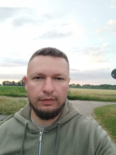 The image is a selfie taken outdoors during daytime. A man with short hair and a beard is the central subject, wearing a green hooded sweatshirt. He is positioned in the foreground, with a background consisting of a field of wheat, trees, and a cloudy sky. The location is identified as Schiffdorf, Germany. The composition is a medium close-up, focusing on the man's face and upper torso.