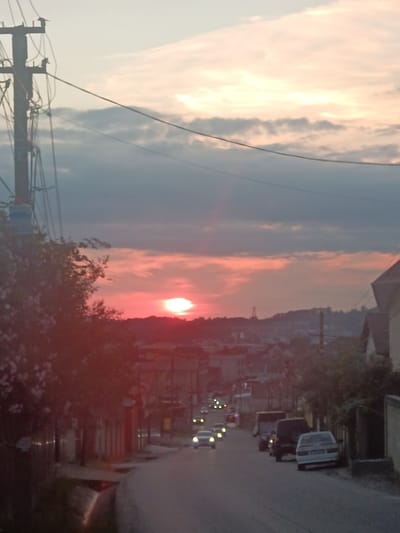 The image depicts a street scene at sunset in Verkhnevesyoloye, Russia. The sky is filled with a mix of dark clouds and hues of pink and orange from the setting sun. A prominent telephone pole with numerous wires is visible on the left side of the frame. The street is lined with buildings and trees, with several cars parked along the roadside and others driving on the street. The overall impression is that of a quiet evening in a residential area.
