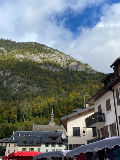 The image displays a village scene in Abondance, France, situated at the base of a large, steep mountain under a partially cloudy sky. The mountain, occupying the upper two-thirds of the frame, is heavily forested with a mix of coniferous and deciduous trees, exhibiting green and yellow autumnal foliage, interspersed with significant grey rock outcrops.
In the mid-ground, the distinctive dark, multi-tiered spire and large dark-tiled roof of Abondance Abbey are visible, with a weather vane at the spire's apex. Surrounding the abbey are traditional alpine-style buildings, characterized by light-colored (white/cream) facades, dark roofs, and numerous windows fitted with dark brown or green wooden shutters. A building on the right features a dark, ornate wooden balcony and an antique-style black street lamp mounted on its exterior wall. A white satellite dish is affixed to a roof in the lower-mid ground, to the left.
The foreground is filled with the upper sections of market stalls, indicated by their large fabric awnings. A prominent red awning is visible on the left, alongside grey and white scalloped awnings extending across the bottom of the frame, obscuring ground-level views, activity, and any potential individuals.