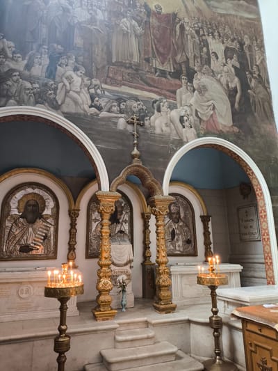 The image shows an interior view of a church or religious building, possibly located in Oktyabrskoye, Russia. In the background, there is a large mural depicting a historical or biblical scene with numerous figures engaged in a water immersion, with several people standing on shore and in the water. In the foreground, there are three arched niches, each containing religious icons adorned with gilded frames and metalwork. Lit candles in ornamental stands are placed in front of the icons. The architecture is ornate, featuring carved pillars and arches decorated with intricate designs. White marble steps lead up to the altar area, where the icons and candles are positioned. Inscriptions in a language other than English are visible on plaques mounted on the walls. The environment appears to be somber and reverential, enhanced by the soft glow of the candlelight.