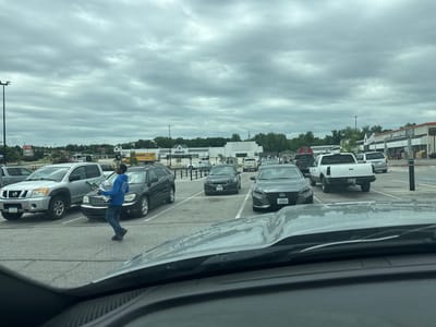 A young Black man in a blue hooded sweatshirt and dark jeans is walking across a paved parking lot. He is carrying a large, flat, rectangular box under his left arm and a smaller, similar-shaped box in his right hand. He is walking away from the viewer and toward a dark gray SUV.
The parking lot is filled with cars and trucks. In the background, there are several buildings, including a Home Depot, a PetSmart, and an "Indoor Lumberyard" with a "Veteran Parking" sign. The sky is overcast with gray clouds. The scene appears to be in Des Moines, Iowa, United States of America, based on the presence of chain stores common in the area.