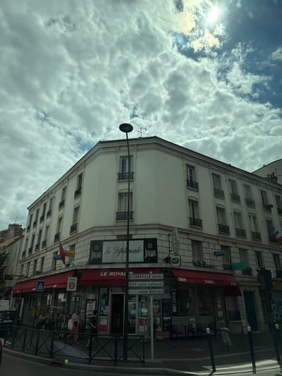 This is a vertical, eye-level, medium shot of a street scene in Alfortville, France, under a cloudy sky with the sun peeking through. The main subject is a multi-story building with a white facade, featuring several windows with dark decorative grilles. On the ground floor, a red awning stretches across the storefront, with "TABAC" and "BAR" clearly visible. Below the awning, there are glass doors and windows revealing merchandise and seating areas, with a few patrons visible inside and outside the establishment.
Prominently displayed signs include "LE ROYAL," "FDJ," "PMU," and "Le Diplômate." A street sign indicates "ALFORTVILLE-CENTRE" and "POC-pôle culturel." A traffic sign reads "ZONE 30," indicating a 30 km/h speed limit zone.
Several people are visible on the sidewalk and near the storefront, some appearing to be customers and others passersby. The overall atmosphere suggests a typical day in a French town, with commercial activity and people going about their routines. The cloudy sky with patches of sunlight indicates the time of day could be late morning or afternoon. The presence of French text on signs confirms the location.