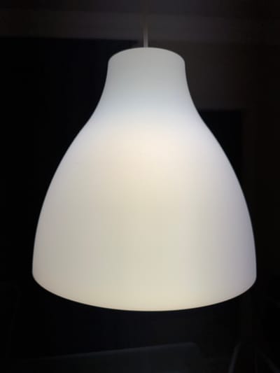 A close-up, head-on shot shows a white, bell-shaped lampshade hanging against a dark background. The lampshade is made of a translucent material that diffuses light, giving it a soft glow. The top of the lampshade is narrower and curves outward to a wider base, creating an elegant silhouette. A thin cord extends from the top of the lampshade, disappearing into the darkness above. The background is uniformly black, with no visible details, emphasizing the lampshade as the sole subject. The image appears to be taken indoors, likely in a room with low ambient light, allowing the lampshade to be illuminated from within. The location of Fañabé, Spain, is not visually represented in this image.