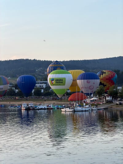This image captures a scenic view of Gródek nad Dunajcem, Poland, featuring a large gathering of hot air balloons ascending over the shimmering waters of Lake Rożnowskie. The balloons, in a variety of vibrant colors and patterns, dominate the sky, with several in mid-flight. A cluster of boats is docked along the shoreline, with many people milling about on the grassy bank and near the water's edge. The background is characterized by lush, tree-covered hillsides, dotted with houses. The overall atmosphere suggests a festive event, likely a hot air balloon festival, taking place during daylight hours.