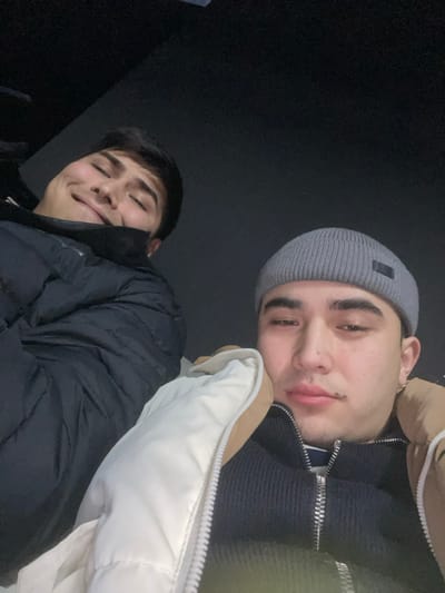 Two young men pose closely for a photo. The first, wearing a gray beanie and a white and tan puffer jacket, is positioned above, looking down with a slight smile. The second man, below, wears a dark puffer jacket and looks upward, also with a gentle smile. The background is a dark, nearly black space.