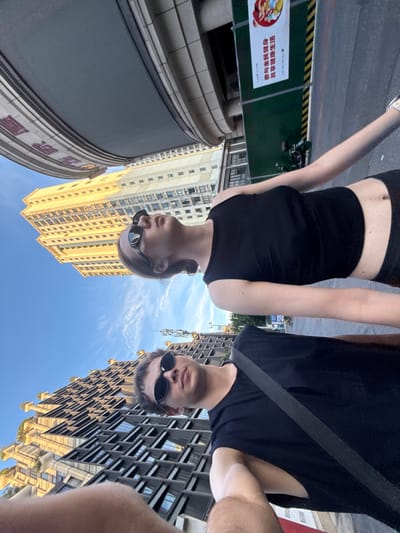 The image is a low-angle selfie featuring two individuals, a man and a woman, looking upwards towards the camera against a backdrop of tall urban buildings and a clear blue sky. Both subjects are wearing sunglasses and dark, casual clothing. The man, on the right, wears a dark t-shirt. The woman, on the left, wears a black sports bra or crop top and black shorts.

The setting is a modern urban environment, likely a street or public square. On the left, a large, curved building with a grey facade and visible purple/red text on its lower section dominates the foreground. Behind the woman, a tall, light yellow high-rise building stands prominently. On the right, behind the man, another striking modern building features a complex geometric facade with many windows and gold-colored accents, reflecting the blue sky.

The weather appears to be bright and sunny, indicative of daytime, with a clear blue sky and strong sunlight illuminating the buildings. A grey paved area is visible at the bottom of the frame, along with a green barrier featuring yellow and black striped tape. Attached to this barrier is a white sign with red Chinese characters. The visible text on the sign reads: "防疫与疾病防治 共享健康生活", which translates to "Epidemic prevention and disease control, share a healthy life." The extreme low angle of the shot emphasizes the height of the surrounding architecture and the expansive sky above the subjects.