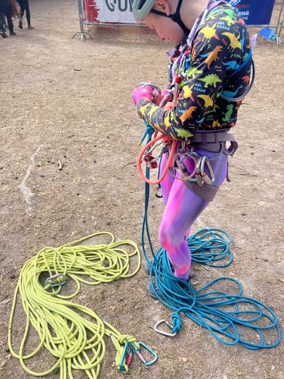 A person, presumably a climber, is shown from a top-down perspective. The individual is equipped with a climbing harness and is dressed in clothing patterned with dinosaurs. A climbing helmet is partially visible on the person's head. The person is surrounded by coiled climbing ropes, one set is blue and another set is yellow-green. The ground is covered in dry grass and dirt. A sign with text "Кий" and a partial view of the city skyline of Lesnoye, Russia is visible in the background.