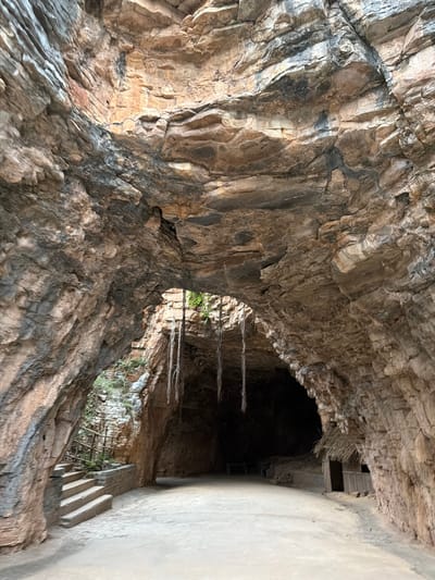 The image shows the entrance to a cave or cavern with rough, textured rock formations arching overhead. The cave floor is composed of compacted earth or sand. To the left, a set of stone stairs with a simple wooden railing leads upwards and out of the cave, with some greenery visible. Hanging down from the cave ceiling are several thick, rope-like vines or roots. To the right, a small, rustic hut constructed of wood and thatch is nestled against the cave wall. The interior of the cave is darker, with some indistinct structures or features visible deeper within. The overall impression is of a natural geological formation adapted for human use or shelter. This is a view of the caves in Gollagutta, India.