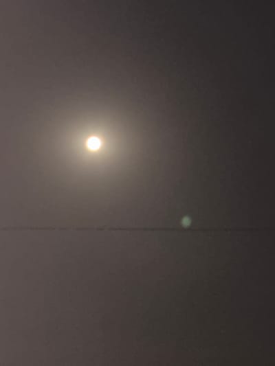 The image depicts a nighttime sky above the city of Slanchevo, Bulgaria. A bright, circular celestial body, likely the moon, is visible in the upper left quadrant of the frame, emitting a soft, diffused glow. Below and to the right of the moon, a smaller, faint green orb is also present, possibly a lens flare or another atmospheric phenomenon. A horizontal line, possibly a power line or a structural element, bisects the lower half of the image, creating a subtle division in the dark, hazy atmosphere. The overall impression is one of a dimly lit, possibly overcast, night.