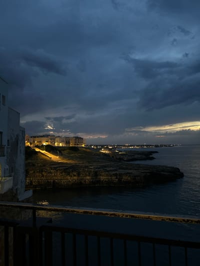 The image captures a twilight scene in Polignano a Mare, Italy. In the foreground, a dark metal railing and a portion of a weathered white building are visible. Beyond these elements, a rugged coastline meets the dark, calm sea. Along the clifftop, a town with illuminated buildings stretches into the distance. A paved pathway, lined with streetlights, ascends a grassy incline towards the illuminated structures. The sky is filled with dramatic, dark clouds, suggesting a stormy or overcast evening. The water reflects some of the distant lights.