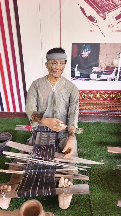 A life-sized mannequin depicting a seated individual engaged in traditional weaving is displayed in Jiyang District, China. The mannequin, with an earthy skin tone, wears a grey headband and a light grey, long-sleeved tunic with visible dark stitching along the collar and sleeves. Its face features black linear markings, possibly representing tattoos or scarification, and similar patterns are visible on its forearms. The mannequin is positioned over a backstrap loom, with its hands holding weaving tools amidst dark and multi-colored threads. A partially woven textile, featuring intricate geometric patterns in hues of red, green, and blue, is visible on the loom. The mannequin sits on a surface covered with artificial green grass. To its left, a dark, woven basket rests on the grass. The background consists of a white wall. On the left side of the background, vertical strips of fabric in varying shades of red and burgundy are displayed. On the right side, two informational posters are affixed. The upper poster illustrates diagrams of weaving tools with Chinese labels. The lower poster displays a photograph of an elderly person, wearing a head covering, also engaged in a weaving process. Below the posters, a patterned fabric with red and yellow geometric motifs is displayed horizontally.