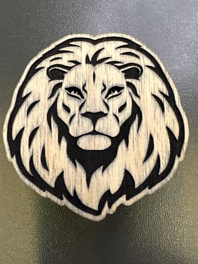 A close-up, top-down shot features a wooden cutout of a lion's head against a dark, textured surface, possibly a table. The lion's mane is depicted with stylized, sharp black lines, contrasting with the lighter, grainy wood. The facial features, including the eyes, nose, and mouth, are also carved with black to define them against the wood. The overall shape of the cutout is oval, with the mane extending outward to create a circular appearance. The background surface is a uniform dark gray with a subtle, uneven texture, suggesting it could be a plastic or coated material. The lighting is even, casting minimal shadows, and highlighting the natural grain of the wood and the precision of the carving. There are no people or animals present in the image. The image is static and devoid of any contextual clues that would place it in Mikhailovka, Russia, or any specific city.