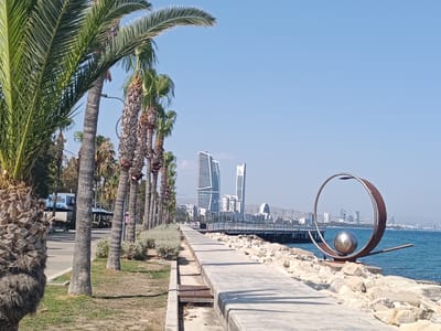This media file depicts a scenic coastal view under a clear, bright sky, suggesting a pleasant daytime with good weather. The foreground is dominated by a paved walkway alongside a grassy area lined with numerous palm trees, their fronds casting shadows.
To the left of the walkway, a street is visible with a building displaying the text "NITRO" in blue. Further down the street, there are awnings and outdoor seating areas, indicating a potential commercial or dining establishment.
The middle ground features the sea, with a pier extending into the water. In the distance, a city skyline is visible, characterized by modern architecture, including several tall, distinctive buildings. A large, abstract metal sculpture with a spherical element is prominently displayed on a rocky embankment to the right of the walkway, overlooking the sea.
The overall scene evokes a sense of tranquility and leisure, likely a popular public space for recreation or sightseeing. While there are no people clearly visible in the frame, the presence of the walkway, pier, and public seating suggests it is a place frequented by visitors. The bright sunlight and clear blue sky indicate it is daytime, likely late morning or early afternoon. The architectural style of the distant city, combined with the palm trees and the coastal setting, hints at a Mediterranean or similar climate.