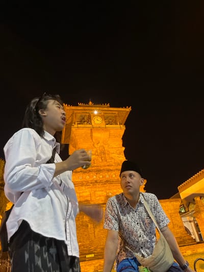 An outdoor nighttime scene in Kauman, Indonesia, features two men positioned in front of a prominently illuminated brick or stone clock tower. The tower, bathed in warm orange light, exhibits multiple tiers and intricate roofline details, with a visible clock face displaying approximately 11:30 or 12:30. The sky above is dark.

To the left, a man with long dark hair and sunglasses perched on his head wears a white long-sleeve collared shirt with dark trim and a dark patterned lower garment. He holds a clear glass containing a yellowish-green liquid in his right hand, which is raised towards his open mouth as if speaking or singing. A dark strap crosses his chest.

To the right, a second man wears a black peci hat, a light-colored short-sleeve patterned shirt, and blue patterned pants. A light-colored shoulder bag is slung across his body. He gazes upwards and to his left, maintaining a neutral expression. His left arm is partially visible, resting out of frame.

Both individuals are in the foreground, with the clock tower dominating the background. A section of another building with a light-colored roofline is visible on the far right.