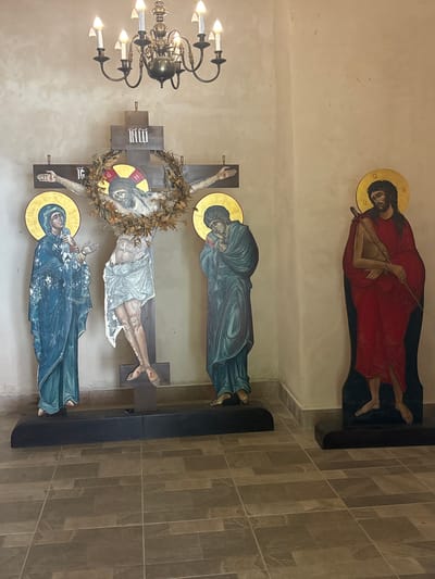 This image depicts an interior scene at the Bachkovo Monastery in Bachkovo, Bulgaria. In the center, a large wooden crucifix is prominent, featuring a painted depiction of Jesus Christ on the cross, adorned with a wreath of leaves. To the left of the crucifix stands a painted figure of Mary, dressed in blue, and to the right, another painted figure, possibly John the Apostle, in a blue robe. Further to the right, a separate, tall, flat painted depiction of Jesus, dressed in red and carrying a staff, is visible. The figures are stylized and appear to be part of religious iconography or artwork. The environment includes a textured wall and a tiled floor. A chandelier hangs from the ceiling.