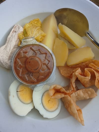 In Gunung Malang, Indonesia, a white plate contains a serving of food. The plate features boiled eggs cut in half, displaying their yellow yolks. Several pieces of boiled potato, cut into wedges, are also present. A small, clear plastic container holds a portion of peanut sauce, its surface textured with a star pattern. To the left of the peanut sauce is a piece of what appears to be steamed tofu, a light beige color. A few leaves of blanched cabbage are visible near the tofu. In the upper right corner, a shiny metal spoon rests on the plate, partly obscuring some of the potato wedges. Scattered around the lower right of the plate are several pieces of fried wonton wrappers, appearing golden brown and crispy. The background is a plain white surface, likely another plate or serving dish.