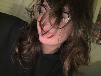 An overhead view looking down at a person with brown hair and black glasses. The person's face is partially obscured by their hair and glasses. The background includes a dark-colored garment, a floor, and part of a table with objects on it. There is no information that this photo was taken in Whitby, Canada.