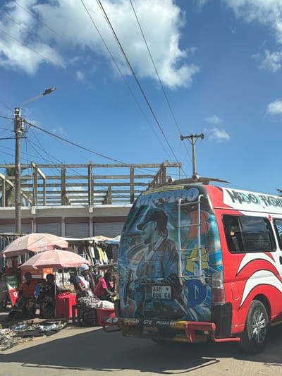 The image depicts a street scene in Pemba, Mozambique. A colorful mini-bus with "MARTICOO" painted on the side and additional decorative graphics is parked or driving on the street. It's partially obstructing the view of a market stall, where individuals are seated under Coca-Cola branded umbrellas. The stalls appear to be selling clothing, with garments hung on display. Litter is scattered on the ground. Overhead, power lines crisscross the sky, connected to utility poles and a street lamp. The sky is blue with scattered clouds. The license plate visible on the bus reads "AAM 008 CA".
