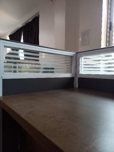 The image depicts an unoccupied interior office environment, specifically an L-shaped cubicle setup. In the foreground, a clean, medium-brown wood-grain desk surface is visible, extending towards the center and right. A set of keys hangs from the underside of the desk on the left. Above the desk, a cubicle partition forms an L-shape, featuring a dark grey lower section and an upper section composed of white-framed panels with translucent horizontal frosted or striped patterns. Behind the partition, light-colored walls, likely off-white, are visible. To the left, through the translucent partition, a window with dark brown or black curtains is partially discernible. To the right, ambient light suggests another window is present, with a faint outline of what appears to be a vertical blind system visible through the translucent panel. A small, light-colored paper document is affixed to the far wall on the right. The ceiling is white. No individuals are present, and no actions or interactions are occurring. The scene is illuminated by natural light. The image provides no visual information to identify the specific city as Oturkpo, Nigeria.