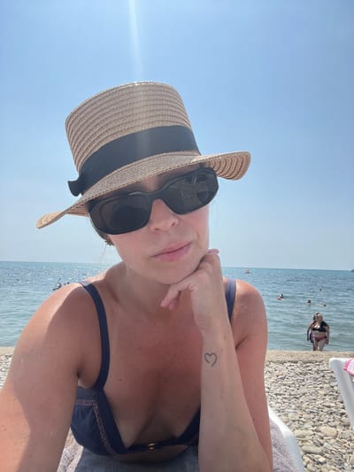 In Blagoveshchenskaya, Russia, a woman is taking a selfie on a sunny day at the beach. She is wearing a straw hat with a black ribbon, black sunglasses, and a dark blue bikini. Her left arm is resting on her chin, and a small heart tattoo is visible on her forearm. The background shows the calm sea with several people swimming and enjoying the water. The beach is covered with pebbles, and lounge chairs are visible in the foreground. The sky is clear and bright blue, with some sun flare from the top right.