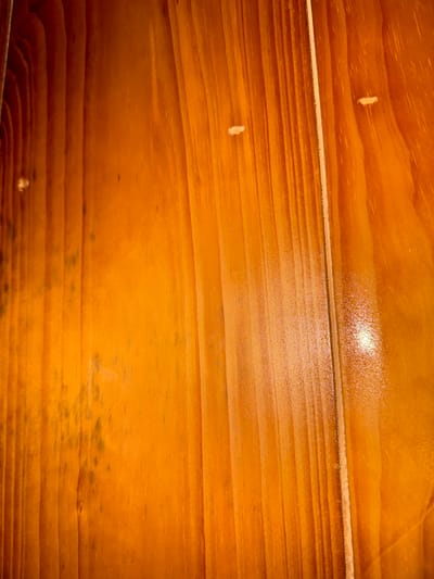 The image shows a close-up of a wooden surface, likely a wall or flooring, made of planks. The wood has a warm, orange-brown color and a glossy finish. The grain of the wood is clearly visible. There are a few small, light-colored spots or imperfections on the surface. In the lower portion of the image, there is a darker area that could be staining or mildew. The top edge of the wooden surface is slightly blurred, and the image appears to be taken in natural lighting. This image has no recognizable connection to Du Già Commune, Vietnam.