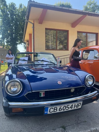 In the city of Skravena, Bulgaria, a blue Fiat convertible is parked outdoors on a paved surface. The car features a chrome bumper, a grille with an Italian flag emblem, and a Bulgarian license plate reading "CB 6054 HP". A bottle of water sits on the hood. A young woman with long dark hair, wearing a black camisole and dark red pants, stands to the right of the car, looking down. To the left, a woman in a white polo shirt and jeans holds a small object. In the background, an orange car is partially visible, and a yellow building with brown-trimmed windows stands behind the vehicles. Trees and a blue sky are also present.