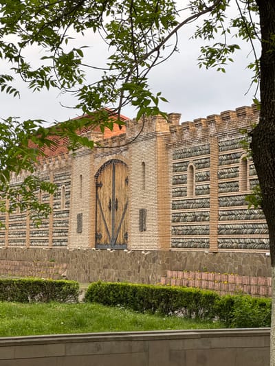 The image captures a section of a building located in Akhieli, Georgia. The structure is composed of brick and stone, with notable decorative brickwork. A wooden door with metal hardware is visible. A portion of a tree with green leaves is in the foreground, partially obscuring the building. Adjacent to the building is a landscaped area featuring greenery and a stone pathway. The sky is overcast.