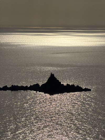 The image depicts a body of water, possibly a lake or sea, reflecting sunlight. A dark, jagged landform, possibly a rocky outcrop or island, is silhouetted against the water. Based on the information provided, it is plausible that the location is Kaluđerac, Montenegro. The foreground shows the reflection of the sun on the water surface. The background is uniformly gray.