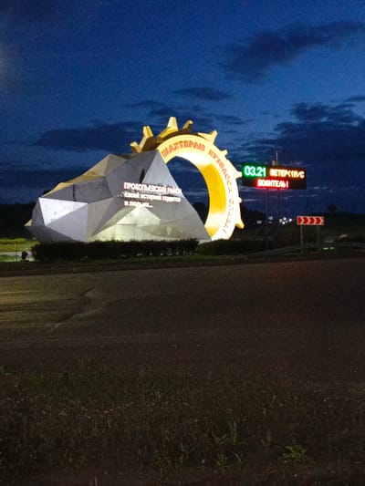 This media file captures a striking nighttime scene in Kalachevo, Russia. The primary subject is a large, stylized monument resembling a gear and a faceted rock formation, illuminated by bright, colorful lights. The monument features prominent text in Russian. On the gear-like portion, the words "ШАХТЕРАН КУЗБАCCA" are visible, translating to "Miner of Kuzbass." The rock formation displays additional text: "ПРОКОПЬЕВСКИЙ РАЙОН своей историей гордится и людьми..." which translates to "Prokopyevsky District is proud of its history and its people..."
To the right of the monument, a digital display board shows the time "03.21" and weather information: "ВЕТЕР < 1M/C" (wind less than 1 meter per second) and "ВОДИТЕЛЬ!" (driver!). This suggests the monument is located near a roadway, possibly as a welcome or landmark for travelers. A red and white directional sign indicating a curve to the right is also visible.
The scene is set against a dark, twilight sky with scattered clouds, indicating it is either early morning or late evening. The ground in the foreground is a dark asphalt road, with a patch of grass and some distant lights suggesting the presence of a town or settlement. The overall mood is somewhat dramatic and impressive due to the large, illuminated monument and the twilight setting. There are no people visible in the image.