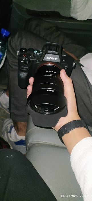 The image depicts an indoor scene in Al-Kiswa, Syria, under low light conditions, captured on 18/10/2025 at 22:29. A black Sony Alpha series mirrorless camera, equipped with a Sony FE 24-70mm lens and a black lens hood, rests on the lap of an individual wearing dark trousers and light-colored shoes. A second individual's right hand, wearing a black digital watch and part of a white long-sleeved garment, is holding the lens of the camera. To the left of the camera, a clear plastic bottle with a blue cap containing clear liquid is partially visible. The background consists of a grey upholstered seat and dark, indistinct areas, consistent with an interior setting such as a vehicle or room.