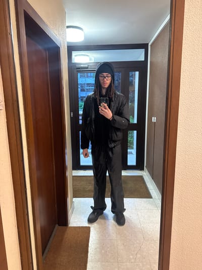 A person wearing a black tracksuit, black beanie, and glasses is standing in front of a mirror, holding a smartphone to take a selfie. The person has dreadlocks. They are in an entryway. The background reveals a view through a window showing greenery and possibly other buildings. The floor of the entryway is tiled. The entryway is located in Les Ulis, France.