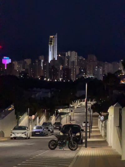 The image depicts a nighttime scene on a paved street in Finestrat, Spain. The street slopes downwards and is lined with streetlights and low walls. Several cars are parked along the side of the street, including a white sedan, a blue hatchback, and a dark SUV. A motorcycle is parked in the foreground. In the background, a dense cityscape with numerous illuminated buildings, including a prominent tall skyscraper, is visible against the dark night sky. One building in the distance displays bright pink and blue lights. Trees and vegetation are present on the slopes leading up to the city.