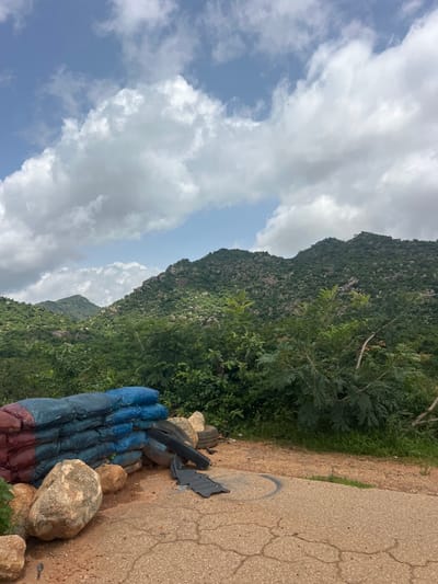 The image shows a roadside scene in Dorowa, Nigeria, featuring a pile of sandbags in blue and red, some large rocks, and discarded tires. A dark, possibly rubber mat-like object is on the ground near the tires. The foreground is a cracked asphalt surface with some gravel and dirt. Behind the sandbags and rocks, lush green vegetation and trees are visible, leading up to a series of rolling, rocky hills covered in dense greenery. The sky above is partly cloudy with patches of blue sky and white cumulus clouds. The overall impression is a natural landscape with man-made debris present.