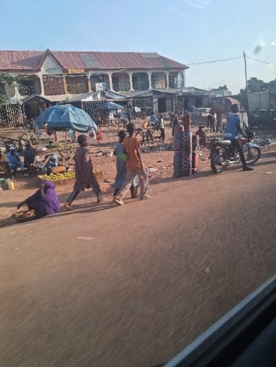 This image captures a vibrant, open-air market scene along a dirt road in Mutum Biyu, Nigeria, under clear daylight. The perspective is from inside a vehicle, indicated by a dark frame in the lower right corner.
In the foreground, a reddish-brown dirt road runs horizontally. On the left side of the road, a person in a purple garment is seated on the ground, attending to a shallow, dark pan containing items. Three individuals walk across the road from left to right: one young person in a brown garment, followed by another in light blue, and a third in an orange shirt and light trousers. To the right, a person in a blue shirt is seated on a red motorcycle, which is parked facing right on the roadside. Nearby, stacks of colorful, patterned cylindrical items are vertically arranged on the ground.
The midground is dominated by a bustling market area situated to the right of the road. This area features multiple makeshift stalls with corrugated metal roofs and at least one prominent blue tarpaulin tent providing shade. Various individuals are visible within the market, sitting, standing, and interacting. Piles of goods, specifically numerous yellow citrus fruits, are displayed on the ground near vendors. The market ground is a reddish-brown dirt surface, scattered with debris.
In the background, a two-story building with a red corrugated metal roof and a light-colored facade stands out. This building features several arched windows or balconies, and solar panels are installed on its roof. A partially legible sign on its facade appears to read "SELIMAZ" or similar. To the right of this building, other less distinct structures are visible. A utility pole with overhead wires is present on the far right. The sky above is clear and light blue.