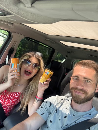 A bright, daytime selfie captures a man and a woman inside a car, likely on a journey through Kaluđerac, Montenegro. The image is taken from a high angle, looking down into the vehicle, and is rotated, with the man's head at the top and the woman's at the bottom.
The man, on the right, has a beard and short hair, and wears a light grey t-shirt adorned with black marine animal patterns including fish, starfish, and squids. He is smiling faintly, looking directly at the camera, with a seatbelt fastened across his chest. His forehead shows some sun glint.
The woman, on the left, has long blonde hair and is wearing a red and white floral top. She has dark sunglasses perched on her head and a wide, open-mouthed expression of excitement or surprise. She wears a silver necklace with a small pendant and beaded bracelets on her left wrist, one of which features a small wing charm. She holds two canned beverages. The can in her right hand is golden-brown with "ICED COFFEE" and "LATTE" clearly visible. The can in her left hand is orange and shows the brand name "E-SCAPE."
The car's interior details include a light-colored headliner, a visible sunroof allowing bright sunlight to flood the cabin, and black leather seats. Green foliage from trees is seen through the car windows, suggesting a natural or scenic outdoor setting. The strong, direct sunlight indicates a clear and bright day, contributing to the cheerful and energetic mood of the subjects.