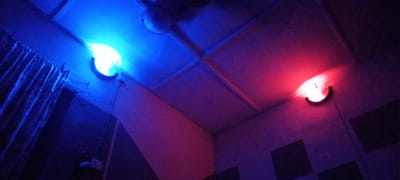 The image shows a low-lit room with two wall-mounted light fixtures. The light fixture on the left emits a bright blue light, while the light fixture on the right emits a red light. The ceiling is composed of square panels, and a ceiling fan is visible in the upper-middle part of the image. A curtain is partially visible on the left. Dark square panels are visible on the lower right wall. The location is not discernible from the image. There are no people or subjects in the image.