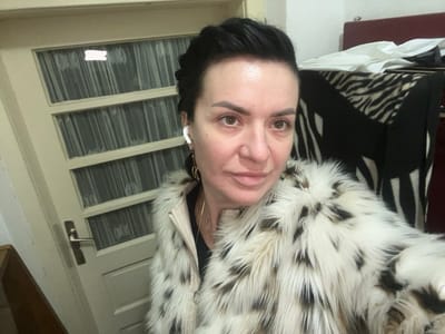 This image depicts an adult female in an indoor setting within Smederevska Palanka Municipality, Serbia. The subject, appearing to be in her 40s or 50s, has dark hair, styled short or swept back. She is looking directly at the camera with a neutral expression, taking a selfie. She wears a multi-toned faux fur coat, primarily white with black and brown markings, over a light-colored jacket and a black garment. A white wireless earbud is visible in her right ear, along with a gold-colored hoop earring and a thin gold-colored necklace.

The background to the left features a light-colored wooden door with multiple rectangular glass panels, each covered by white sheer curtains or vertical blinds. A silver-toned door handle is visible. To the right, a piece of furniture, possibly a chair or headboard, has a visible dark red/maroon section at its top edge. A black and white zebra-print blanket or cover is draped over the lower part of this furniture. The overall lighting is consistent with an indoor environment.