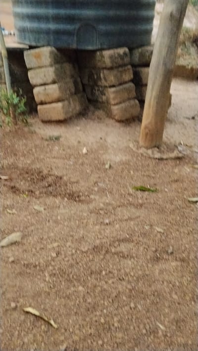 The scene, located in Isieke, Nigeria, displays an outdoor water storage setup. A large, dark grey, horizontally ribbed water tank is elevated, supported by a structure of stacked rectangular concrete blocks. Four blocks are visibly stacked vertically on the left, forming one support pillar, while additional blocks comprise a second support section to the right, partially obscured by a thick, light brown wooden post or tree trunk. A slender, lighter-colored vertical pole stands to the far left of the tank support, with a small patch of green vegetation at its base. The ground consists of reddish-brown, loose earth with scattered small pebbles and dry organic matter. No individuals, actions, or specific interactions are present in the image.
