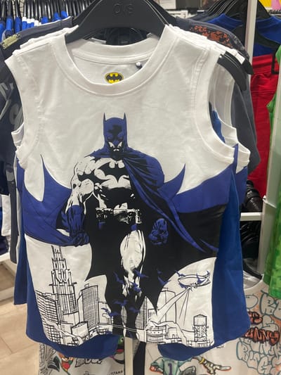A white sleeveless t-shirt with a graphic of Batman is displayed on a clothing rack. The t-shirt features a large illustration of Batman in a dynamic pose, with a cityscape and a blimp in the background. The Batman logo is visible on the upper chest area of the shirt. The t-shirt is hung among other clothing items on black hangers. The scene is indoors, likely in a retail store. There is no indication of Vallo della Lucania, Italy in the image.