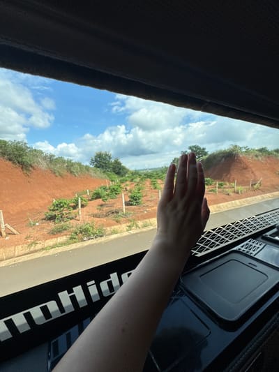 The image shows a person's left hand, palm facing forward, held up to a window. The hand is in the foreground, obscuring part of the view. Through the window, a landscape is visible, featuring a red, hilly terrain with scattered green vegetation and small plants planted in rows. The sky is blue with scattered white clouds. Below the window, a black surface with a white logo that appears to read "toHide" is visible. There is also a textured black grid pattern and a dark rectangular object on this surface. The scene suggests the person is inside a vehicle, possibly a truck, looking out at a rural area. The location is identified as Pleiku, Vietnam.