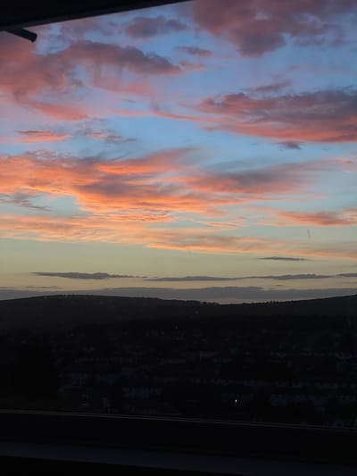 The image depicts a sunset over a residential area in Brighton, United Kingdom. The sky is filled with dramatic clouds, painted in hues of pink, orange, and blue, indicating the sun is low on the horizon. Below the sky, a dark silhouette of hills and houses forms the foreground. The houses appear to be densely packed, with rooftops visible against the darkening landscape. The overall impression is one of a serene, yet potentially moody, evening scene. No people or specific actions are discernible in the image.